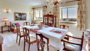A dining area with a table set for four, wooden chairs, a chandelier, and a view through large windows. - Inn the Beach Apartments (Albufeira)