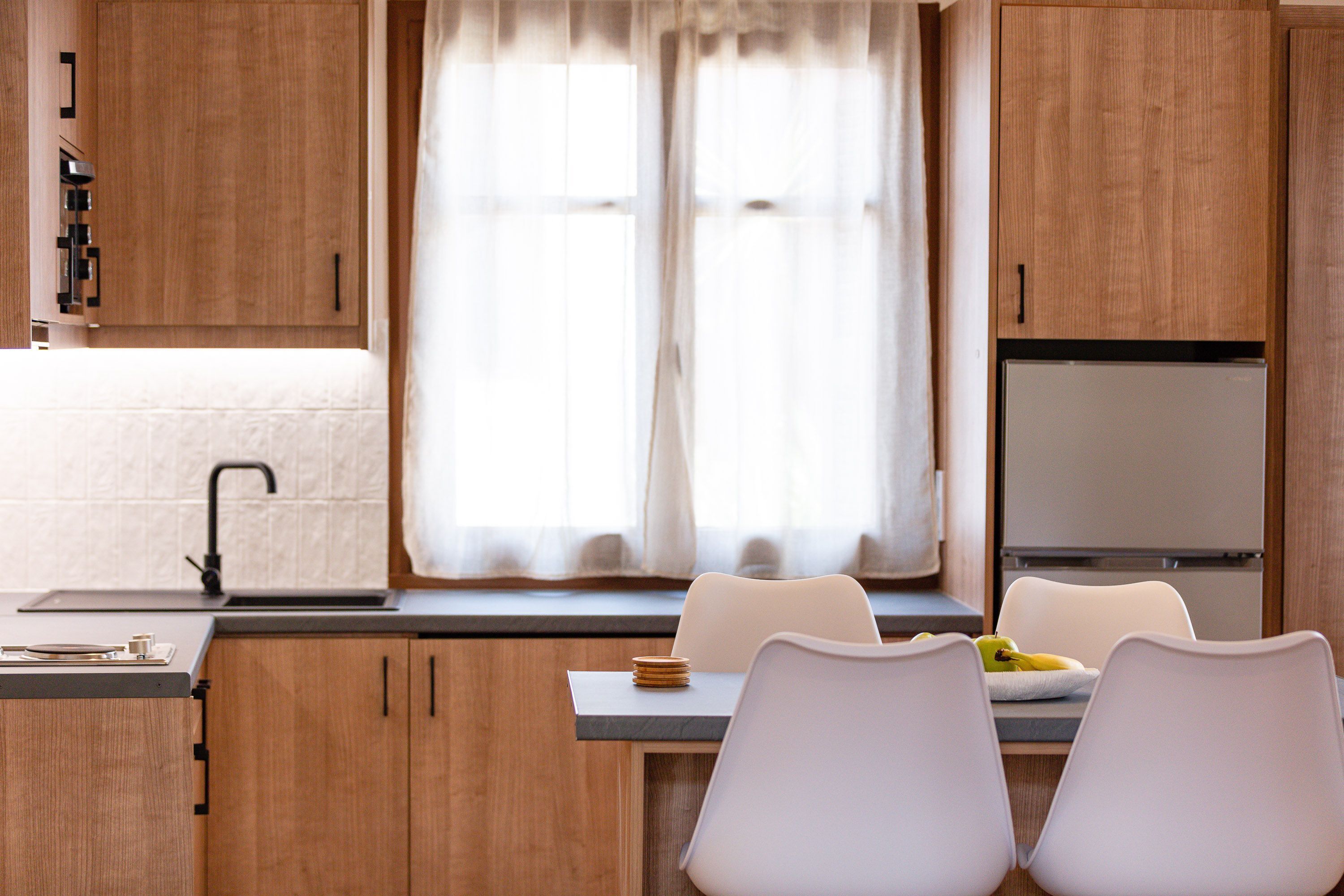 Une cuisine moderne avec des meubles en bois, un réfrigérateur encastré, une fenêtre ornée de rideaux légers et un coin repas comprenant deux chaises blanches et une table.