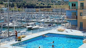 A marina with numerous boats docked, a swimming pool with people, and a building with balconies.