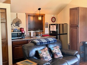 A kitchen with a microwave, coffee maker, and refrigerator. There is a dining area with a leather couch and pillows.