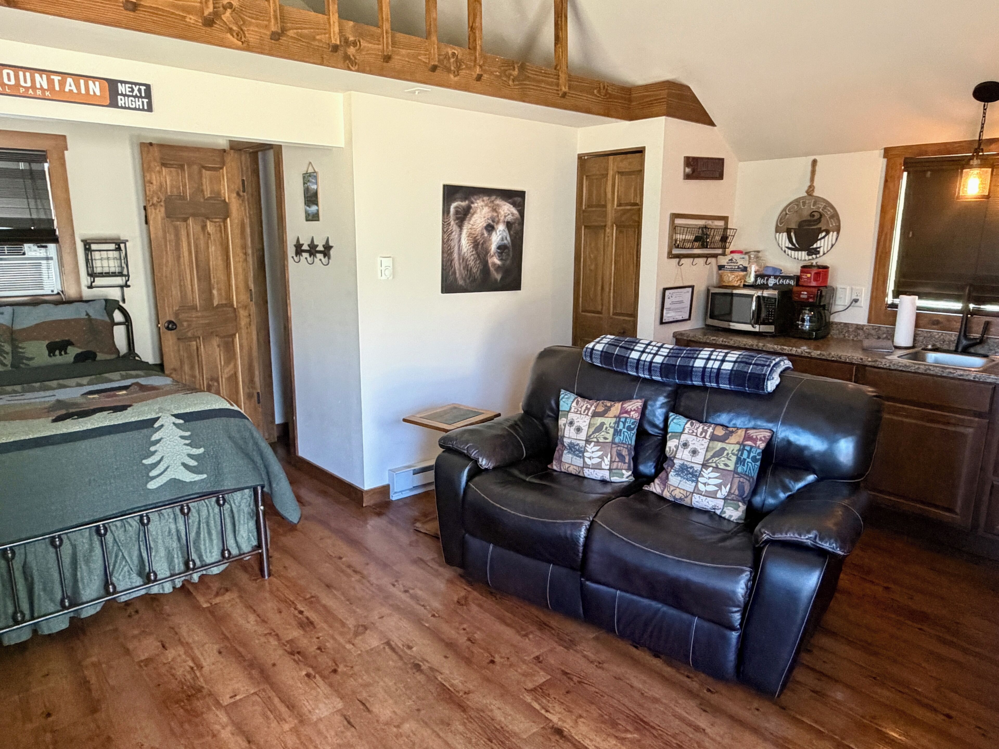 A cozy room with a bed, a leather sofa, a wooden floor, a kitchen area, and a sign indicating a mountain park.