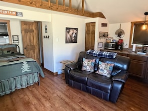 A cozy room with a bed, a leather sofa, a wooden floor, a kitchen area, and a sign indicating a mountain park.