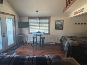 Une piÚce comprenant une table à manger, deux tabourets de bar, un ventilateur de plafond, un téléviseur fixé au mur, un tableau encadré et un lit avec une courtepointe.