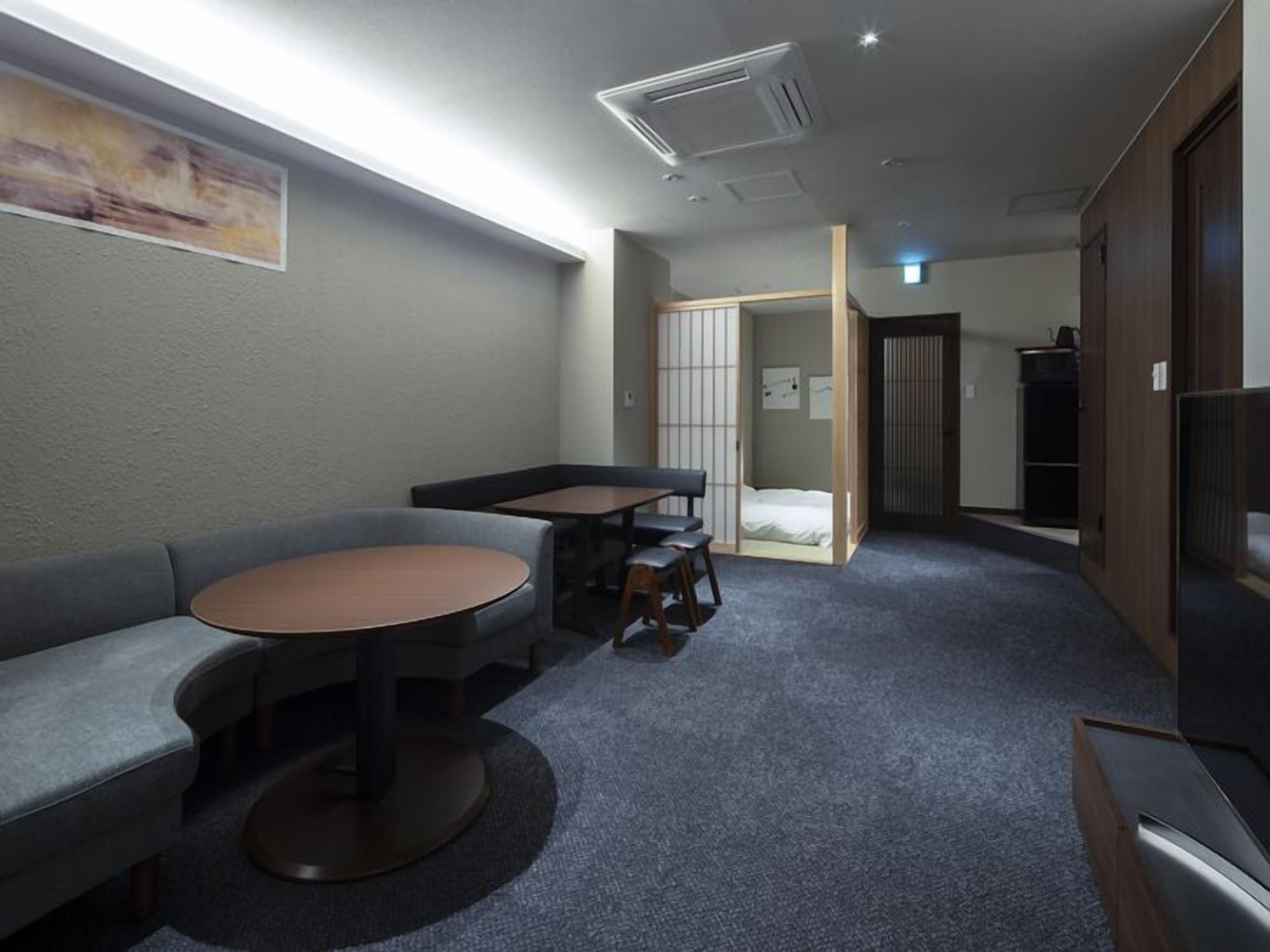 A compact hotel room with a sofa, a small round table, a cabinet, and a sliding door leading to a bed.