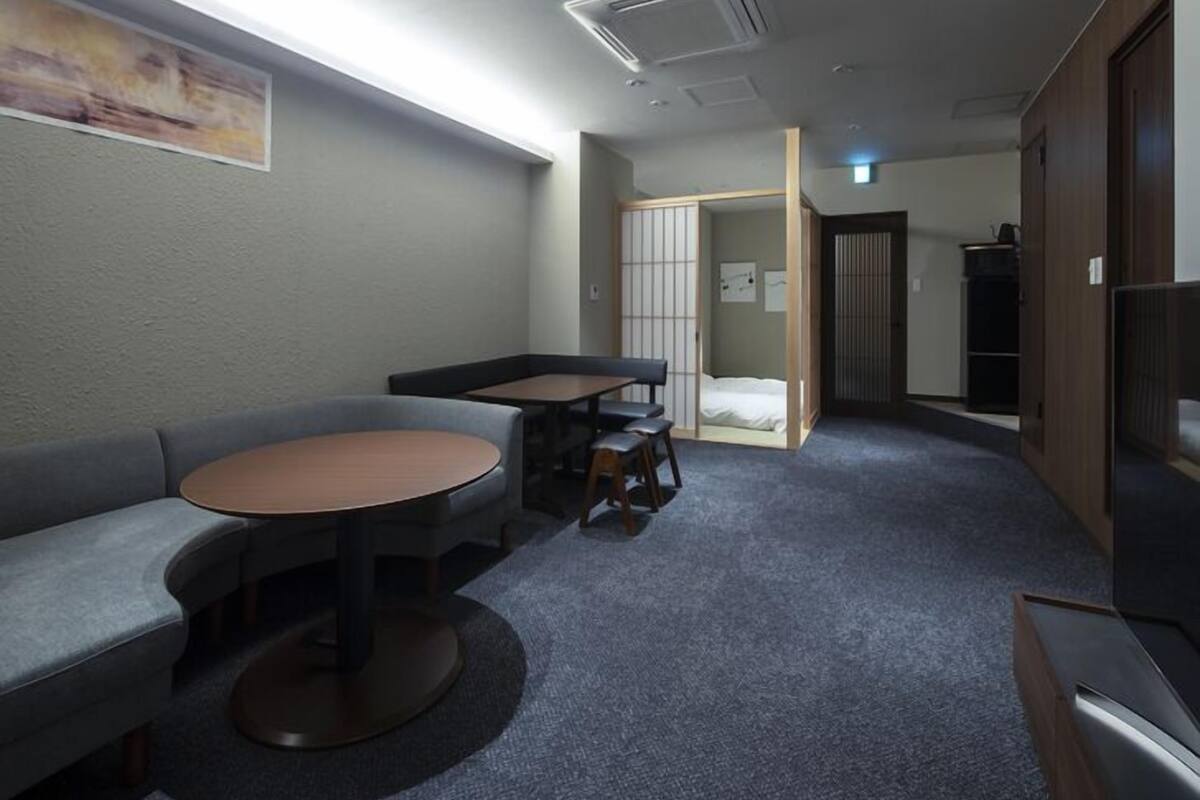 A compact hotel room with a sofa, a small round table, a cabinet, and a sliding door leading to a bed.