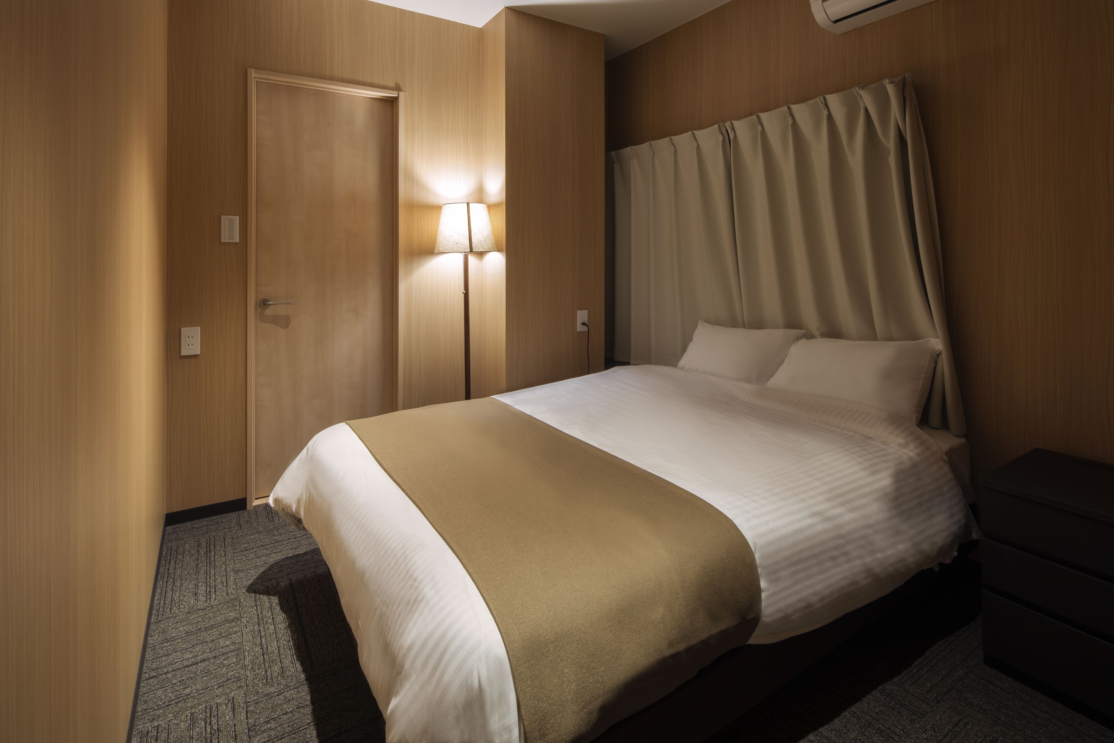 A hotel room with a bed, a bedside table, a lamp, and a door.