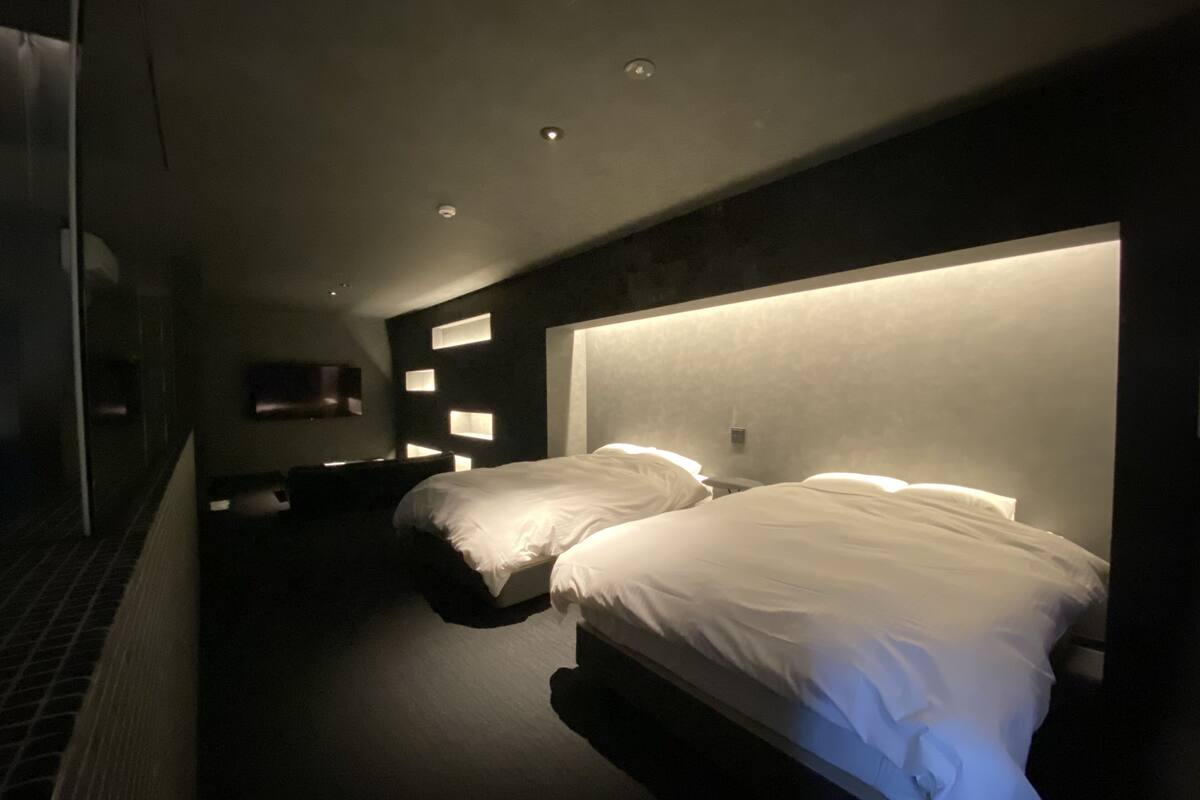 A modern hotel room with two beds, recessed ceiling lighting, and a dark color scheme.