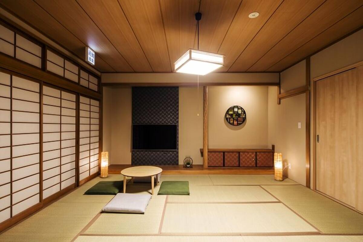 A traditional Japanese room with tatami flooring, a low table, cushions, a shoji screen, a tatami mat wall, a hanging scroll, and a wooden cabinet.