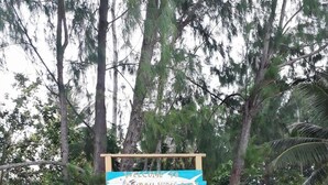A wooden pathway leading to a sign that reads "Welcome to Andau Hars Stai" with a backdrop of trees and a sandy area. - Andau Homestay (Saprokren)