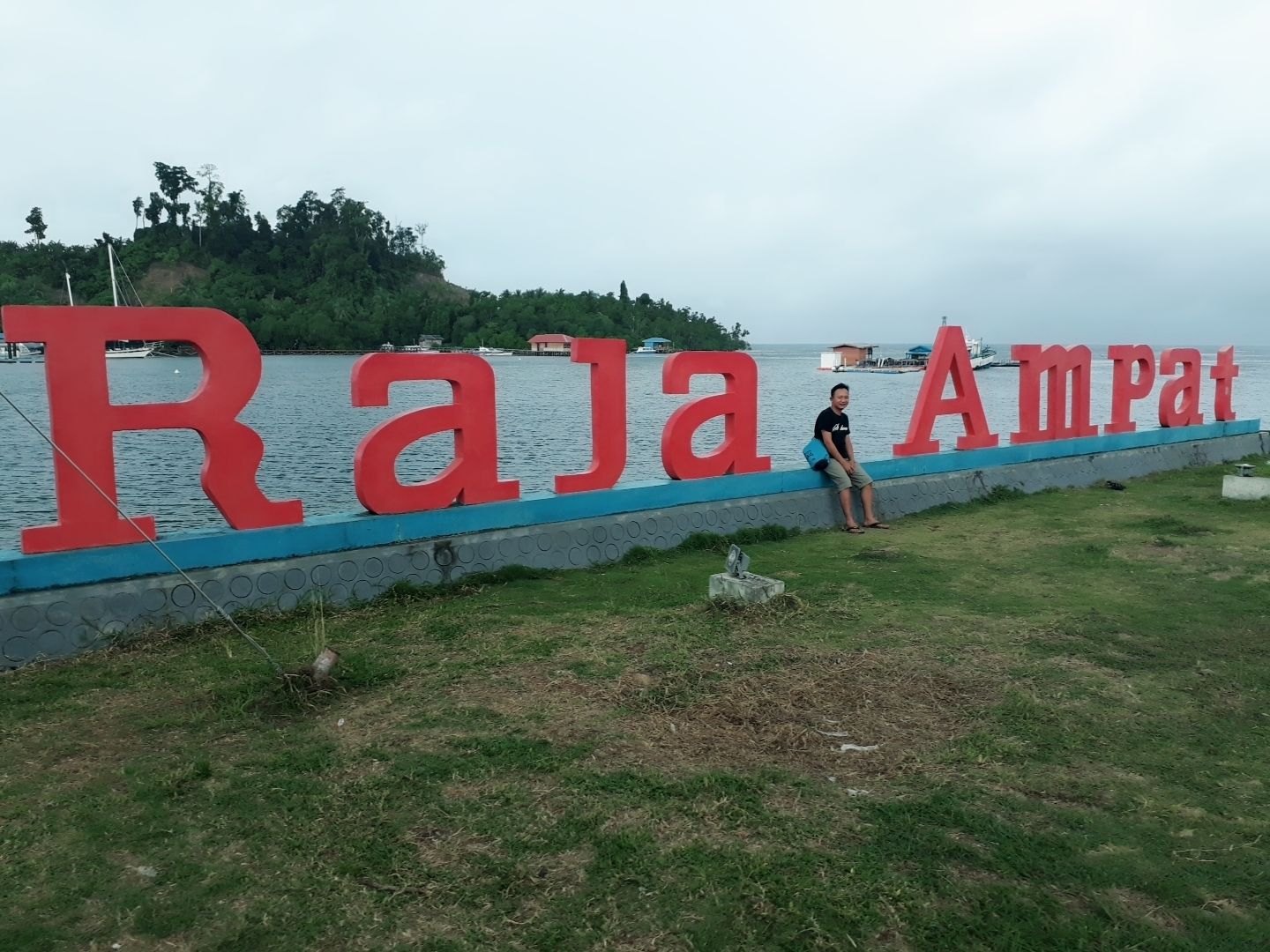 A sign with the text "Raja Ampat" in large red letters, a grassy area, and a body of water with boats in the background.