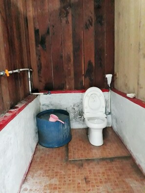 A small, narrow bathroom with a wooden partition, a white toilet, and a blue bucket. - Andau Homestay (Saprokren)