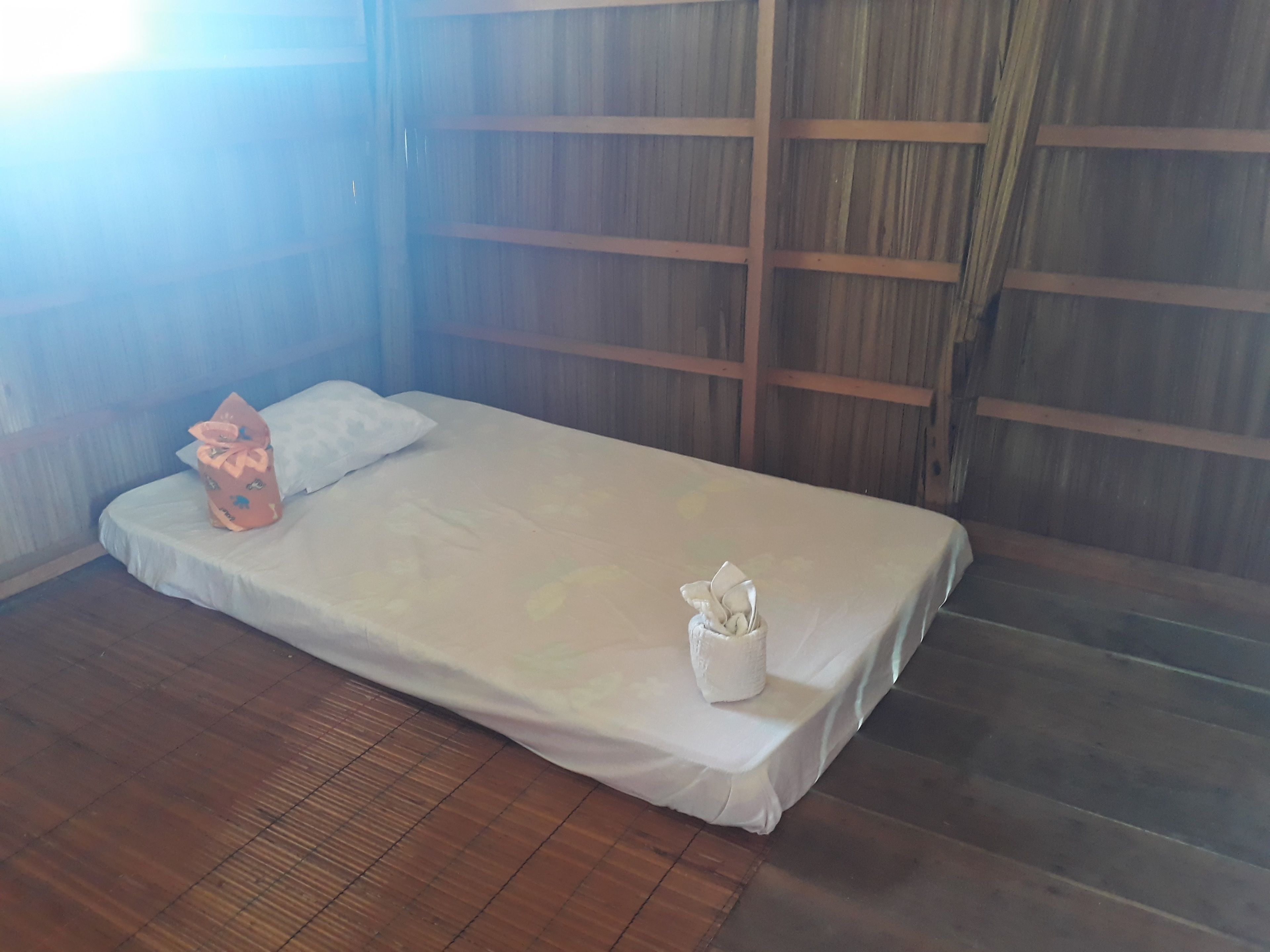 Uma cama de solteiro com colchão e travesseiro brancos, um pequeno recipiente branco e uma sacola de presente rosa no chão.