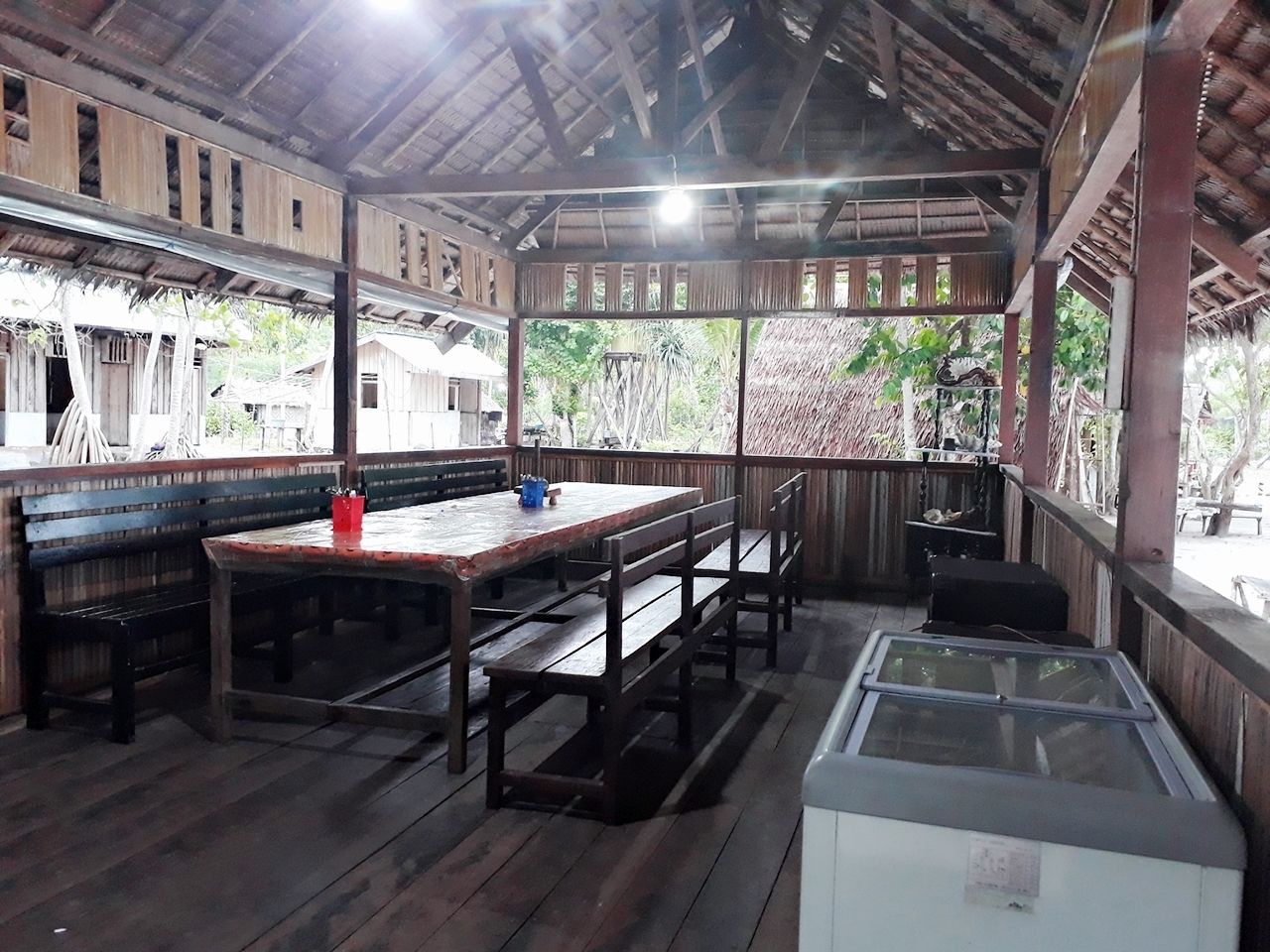 Un comedor de madera con una mesa, bancos, un refrigerador y una estructura de techo de paja.