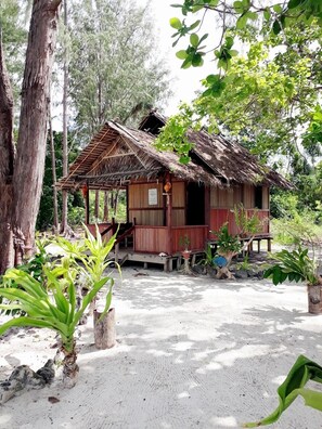 A rustic wooden house with a thatched roof, surrounded by lush greenery and a sandy ground. - Andau Homestay (Saprokren)