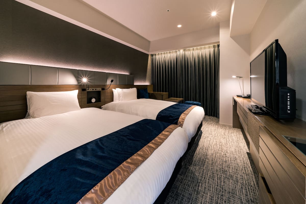 A hotel room with two beds, a TV, a desk, and a large window with curtains.