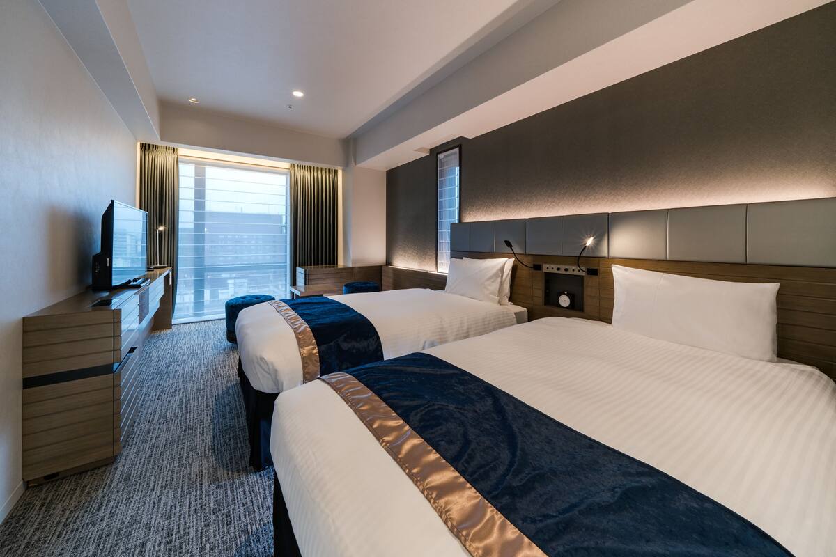 A hotel room with two beds, a TV, a desk, and a large window with curtains.