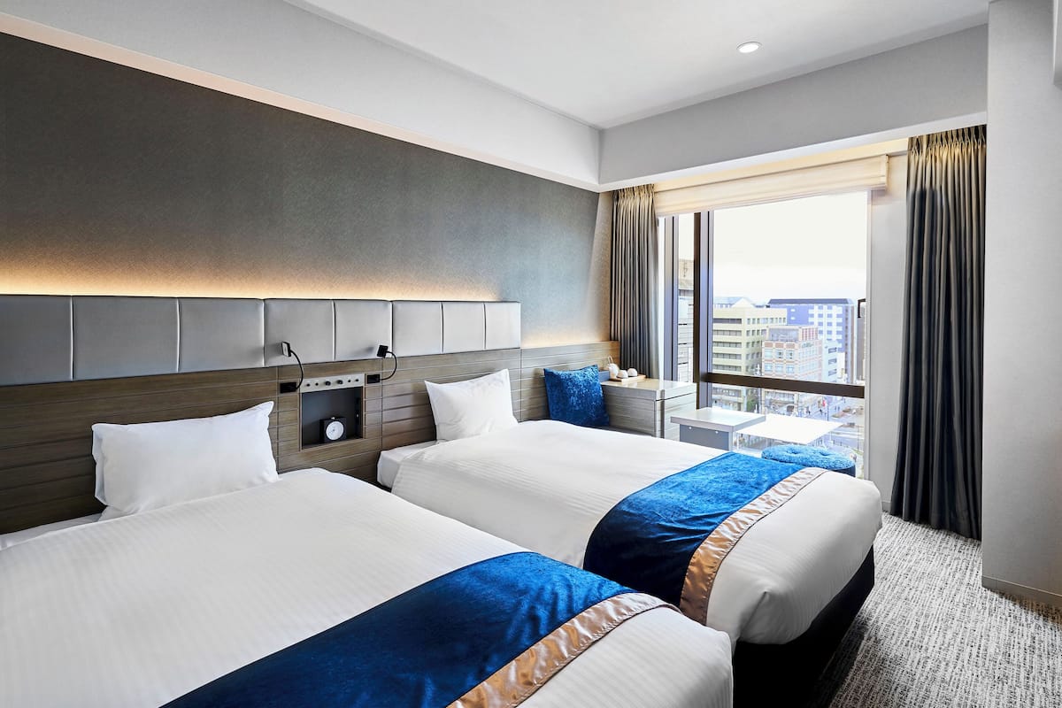 A hotel room with two beds, a nightstand, a window with curtains, and a view of the city.