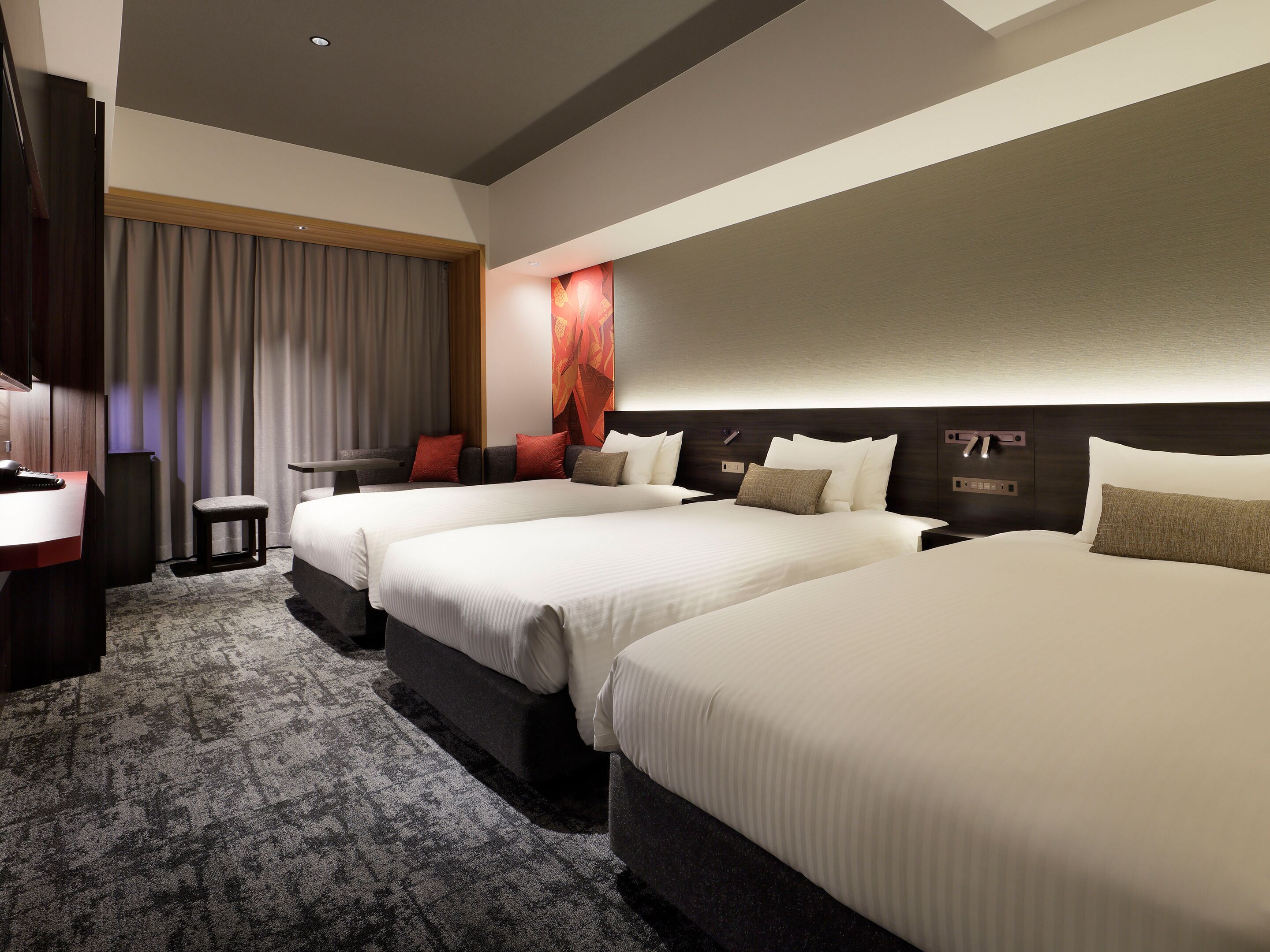 A hotel room with three beds, a desk, a chair, a small table, and a large window with curtains.