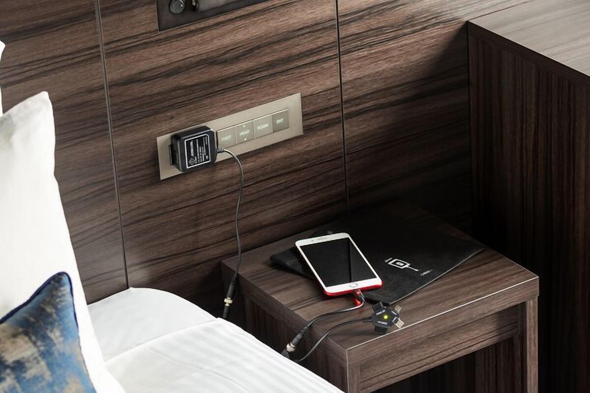Una cama bien hecha con una almohada, una mesita de noche con un teléfono inteligente y cargador, y un enchufe en la pared.