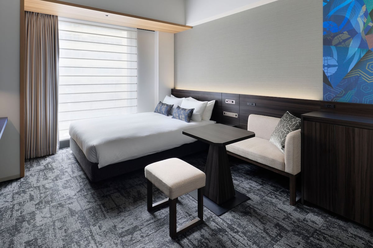 Una habitación de hotel con una cama, un escritorio, una silla, un banco, una ventana con cortinas y un cuadro en la pared.