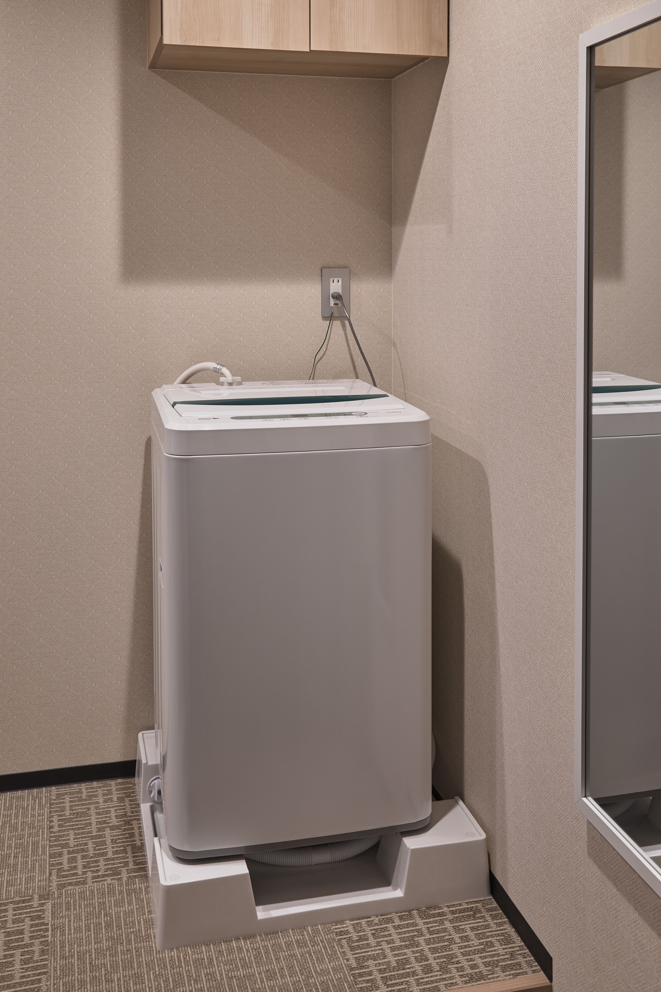 Une petite machine à laver blanche, fixée au mur dans un coin, avec un meuble en bois au-dessus.