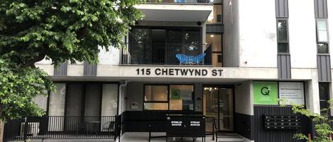 A modern building with a sign reading "115 CHETWYND ST" and a balcony with blue chairs.