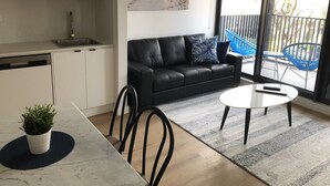 A modern kitchen with white cabinets, a black leather sofa, a dining table with chairs, and a large window with a view of a balcony. - Q Squared Serviced Apartments (North Melbourne)