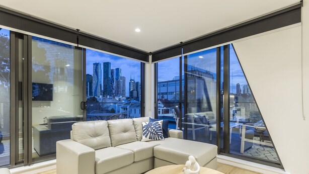 A modern living room with a large window offering a city view, a beige sectional sofa, and a round coffee table with a decorative item. - Q Squared Serviced Apartments (North Melbourne)