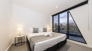 A modern bedroom with a large bed, two bedside lamps, and a city view through a large window. - Q Squared Serviced Apartments (North Melbourne)