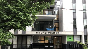 A modern building with a sign reading "115 CHETWYND ST" and a balcony with blue chairs. - Q Squared Serviced Apartments (North Melbourne)