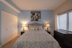 A bedroom with a patterned bedspread, two bedside lamps, a headboard, a painting above the bed, a window with blinds, and a dresser.