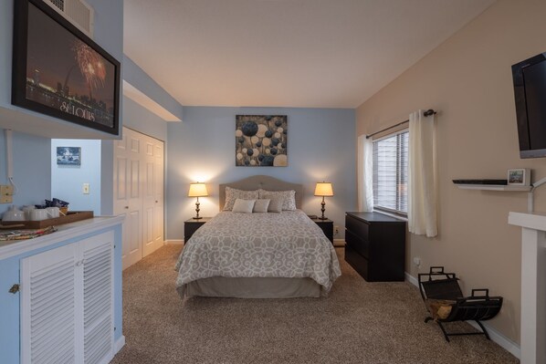 A bedroom with a bed, two bedside tables, a nightstand, a dresser, a window with curtains, and a wall-mounted TV.