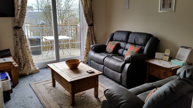 A living room with a leather sofa, a wooden coffee table, and a view through a glass door to an outdoor area with a wooden deck and chairs. - Apartment 4, Skye Holiday Apartments, Phoenix Flats (Portree)