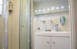 A modern bathroom with a glass shower enclosure, a white sink with a mirror above, and a wall-mounted soap dispenser.