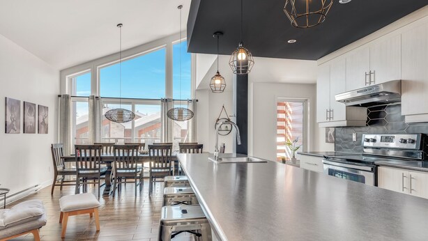 A modern kitchen with a large island, stainless steel appliances, and a dining area with wooden chairs and a table. - Compfortable Chalet to Rent at Stoneham located at the Mountain (CITQ: 287663) (Stoneham-et-Tewkesbury)