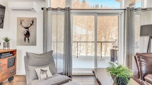 A modern living room with a grey sofa, a wooden side table with a plant, and a large window with curtains. - Compfortable Chalet to Rent at Stoneham located at the Mountain (CITQ: 287663) (Stoneham-et-Tewkesbury)