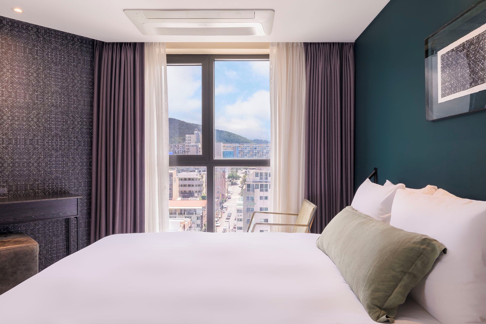 Une chambre d’hôtel avec un grand lit, une vue sur la ville par la fenêtre, un mur texturé de couleur sombre et un luminaire au plafond.