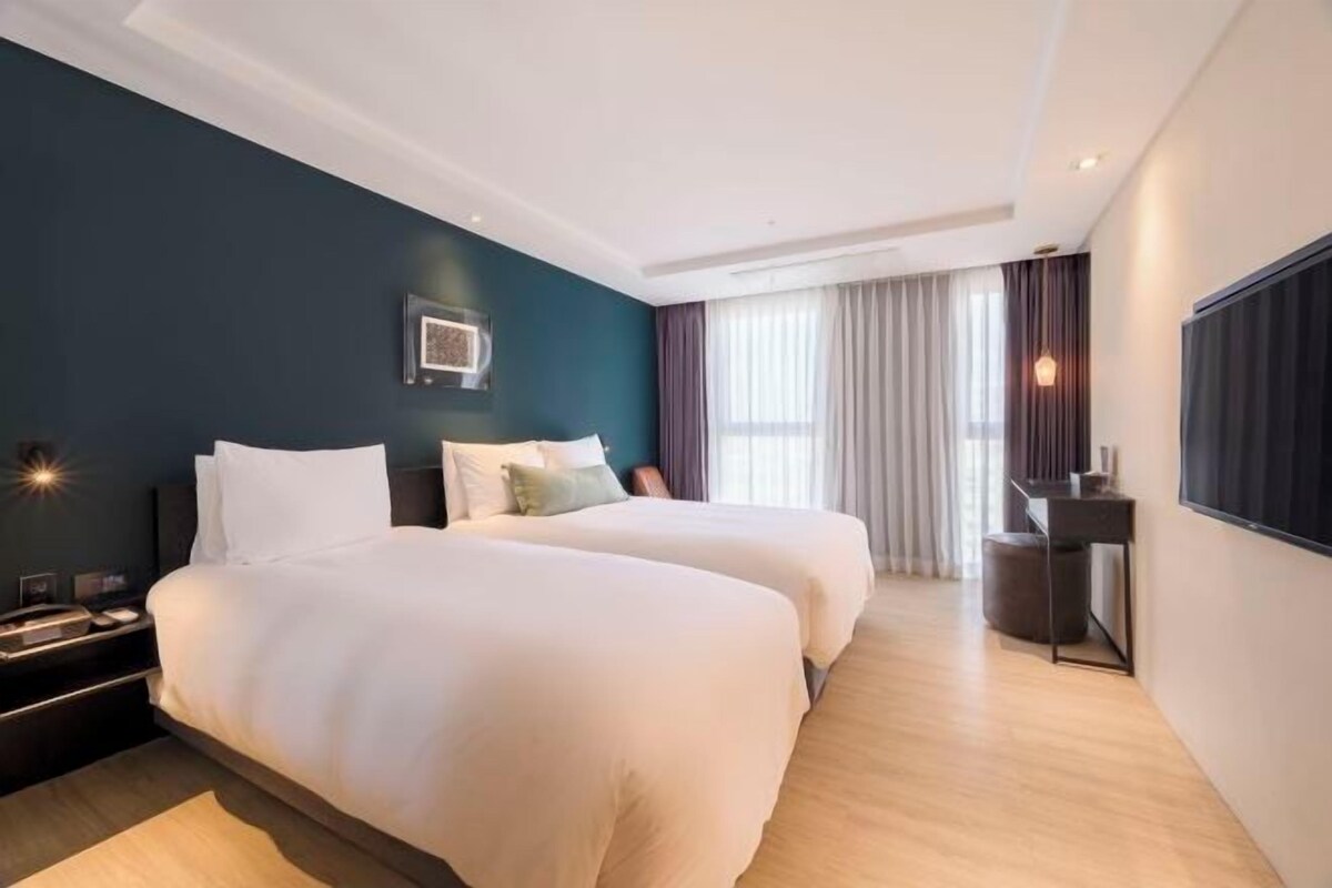 Une chambre d’hôtel avec deux lits, un bureau, une chaise et une grande fenêtre avec des rideaux.