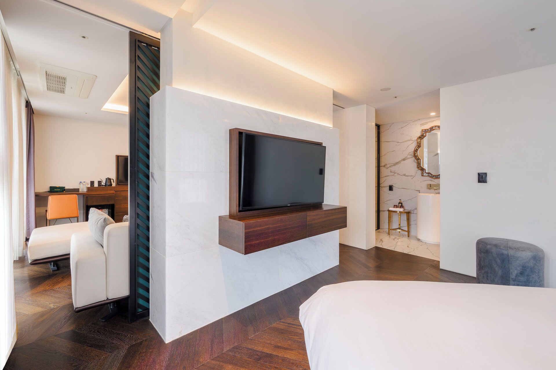 A modern hotel room with a flat-screen TV mounted on a wall, a bed, a seating area with a chair, and a bathroom with a marble wall and a mirror.