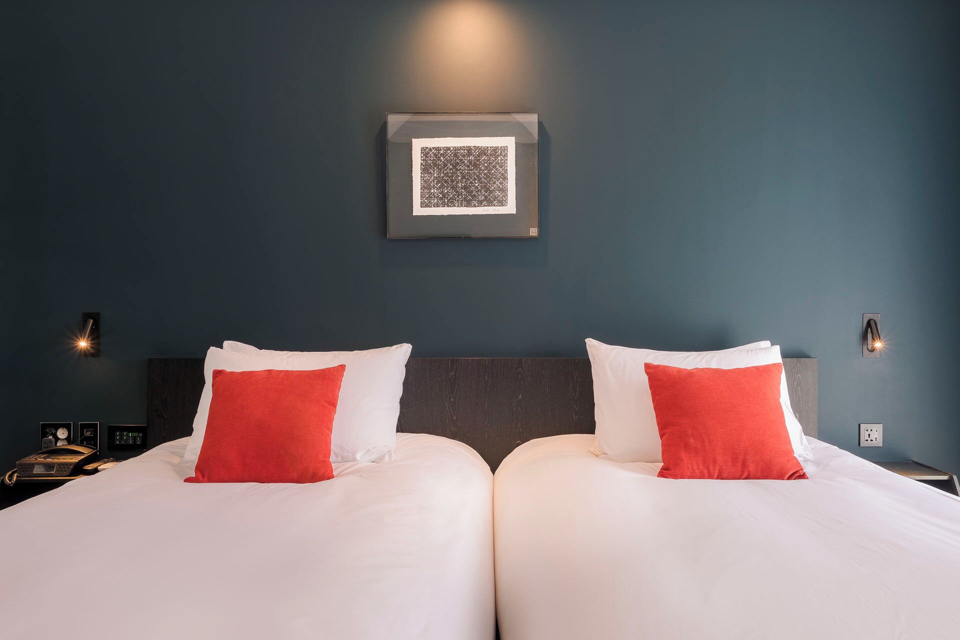 A hotel room with two beds, each with white bedding and red pillows, a dark blue wall, and a framed picture above the beds.