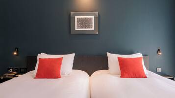 A hotel room with two beds, each with white bedding and red pillows, a dark blue wall, and a framed picture above the beds.