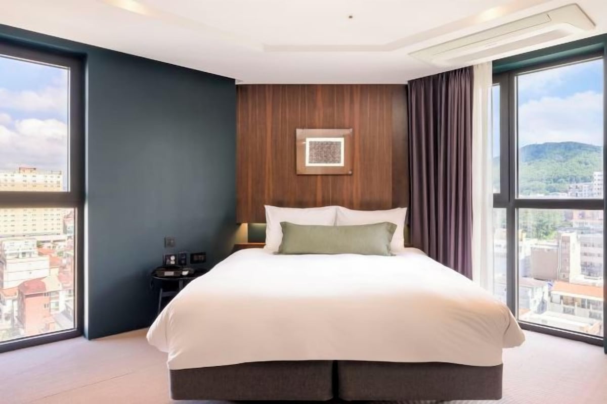 A hotel room with a large bed, a nightstand, a wall-mounted picture, and a view of the city through large windows.