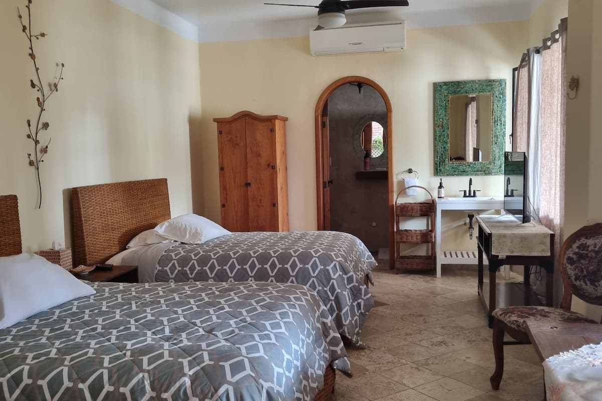 A bedroom with two beds, a wooden wardrobe, a bathroom with a sink and mirror, and a ceiling fan.