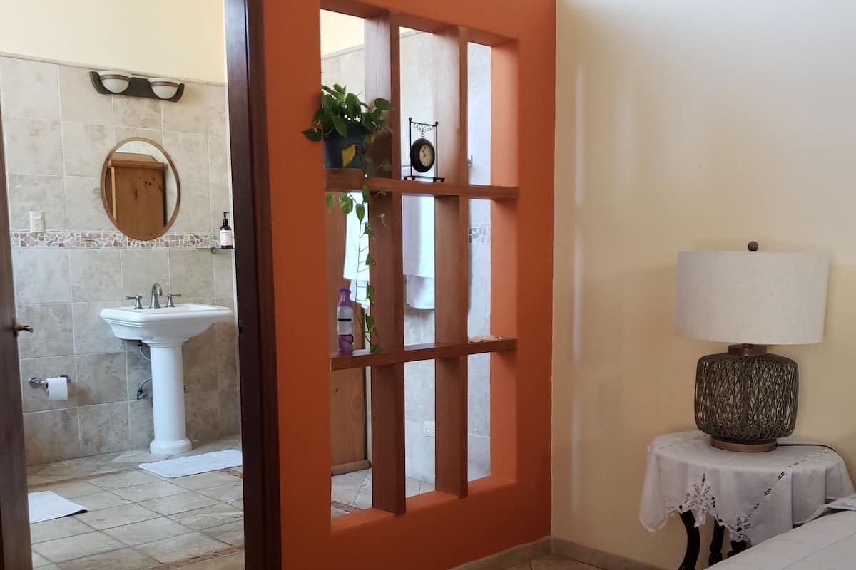 A bedroom with a bed, a nightstand with a lamp, and a view of a bathroom with a sink and mirror.