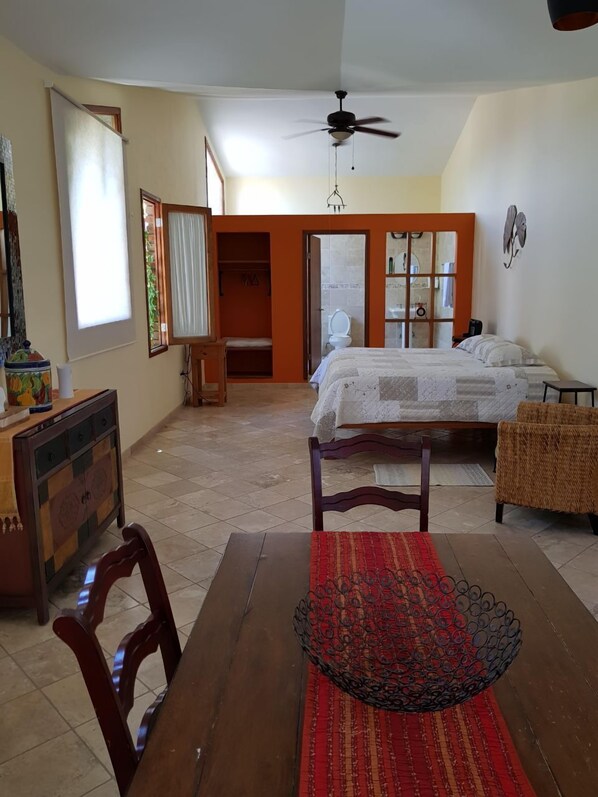 Une chambre à coucher avec un lit, un ventilateur de plafond, une armoire et une salle de bain visible à travers une porte ouverte.