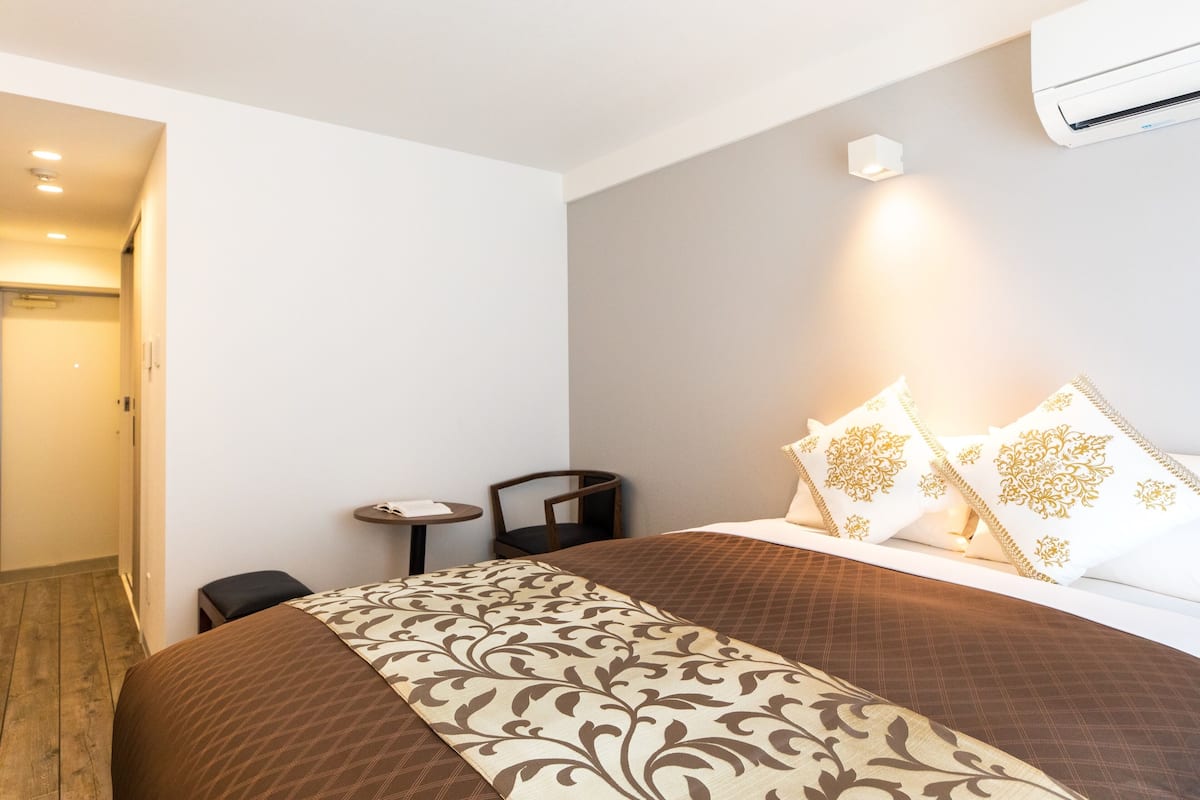 Quarto de hotel com cama, uma pequena mesa, uma cadeira e unidade de ar condicionado.