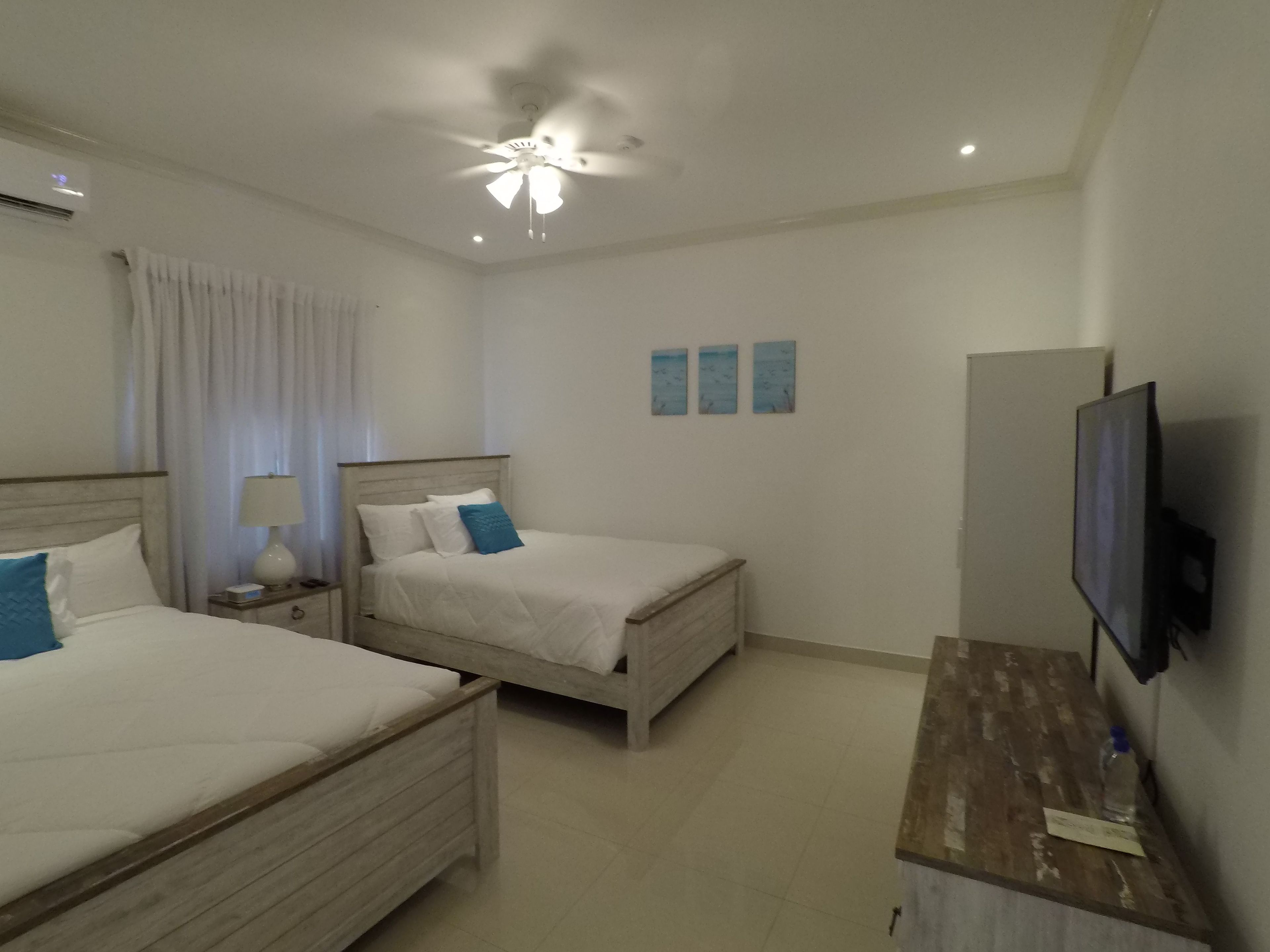 Une chambre d’hôtel avec deux lits, un ventilateur de plafond, un téléviseur à écran plat et une table de chevet en bois.