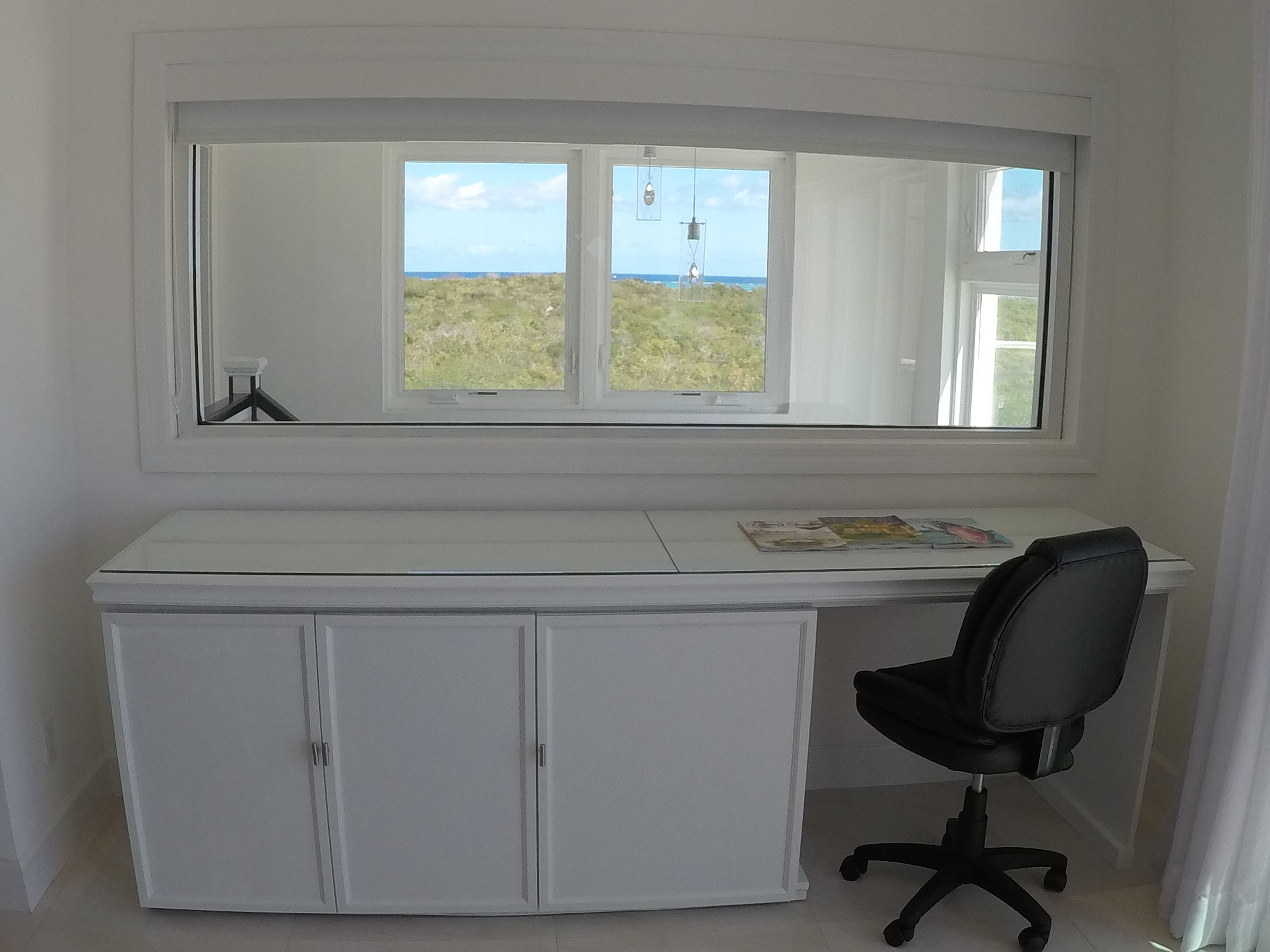 A white desk with a large mirror above it, a black office chair, and a view of the outdoors through the window.