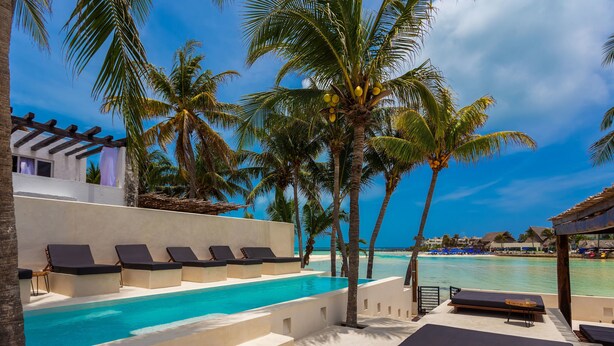 Un espace aménagé au bord de la piscine, avec des chaises longues, une petite piscine, des palmiers et une vue sur l’océan.