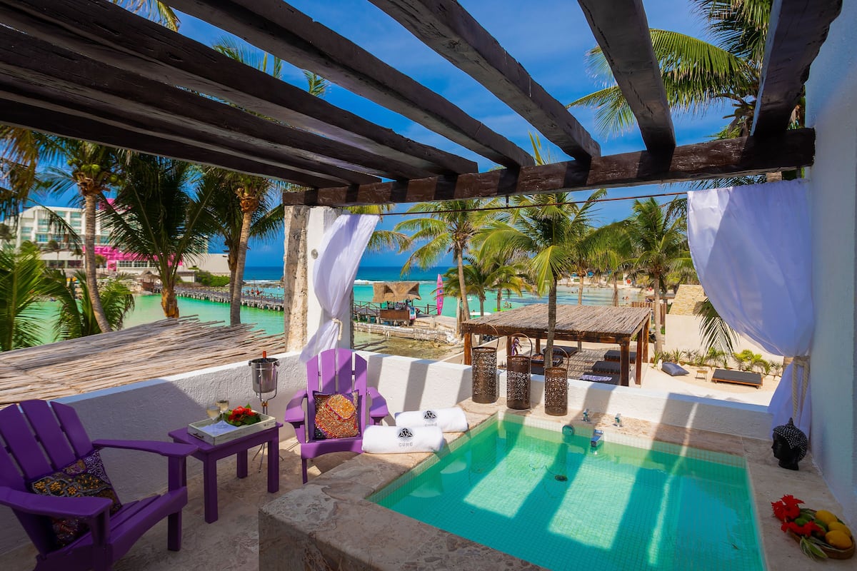 Un espace piscine avec vue sur l’océan, une pergola en bois, des chaises violettes et une petite table avec une boisson et des fleurs.